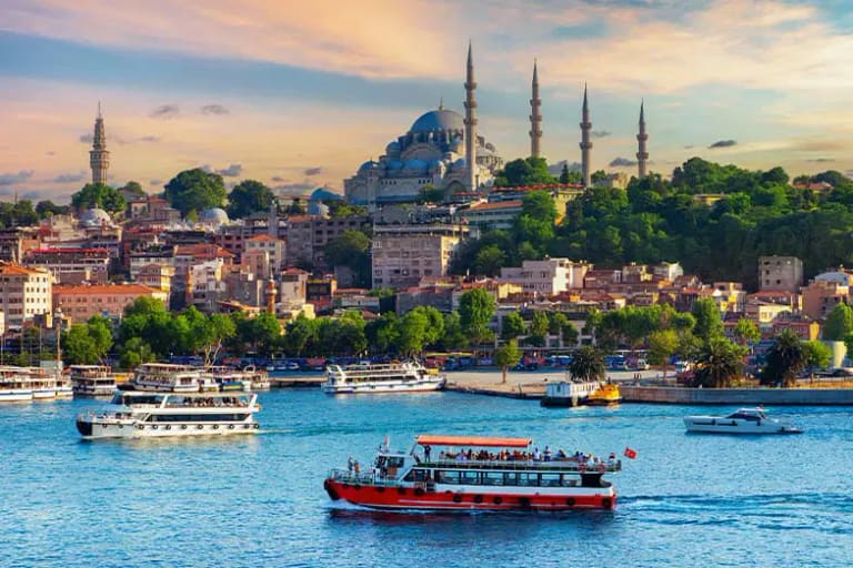 Des ferries traversant l’eau au coucher du soleil, avec une mosquée et ses minarets sur la colline au-dessus de la ville